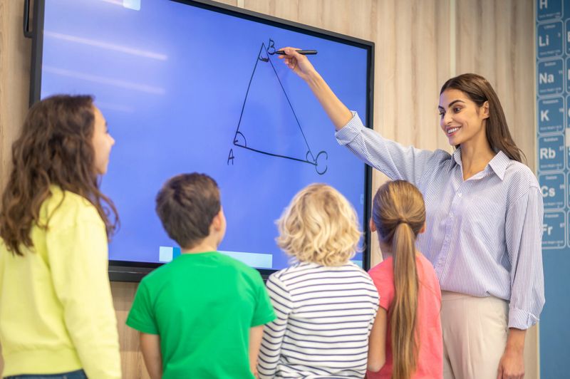 Interesting geometry. Young smiling woman drawing showing triangle on blackboard standing together with kids in class during lesson