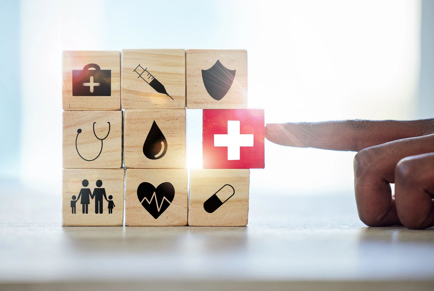 Wooden blocks with healthcare icons and a hand pointing at a red block with a white cross.