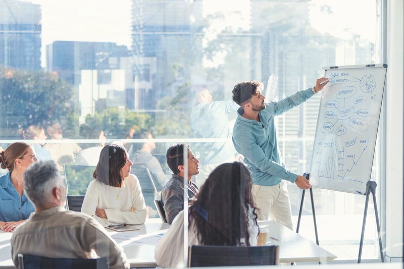 Business people watching a presentation on the whiteboard. A man is writing on the whiteboard with charts and graphs. They are sitting in a board room, there are laptop computers and technology on the table. All are casually dressed. There is a window behind him with city views.