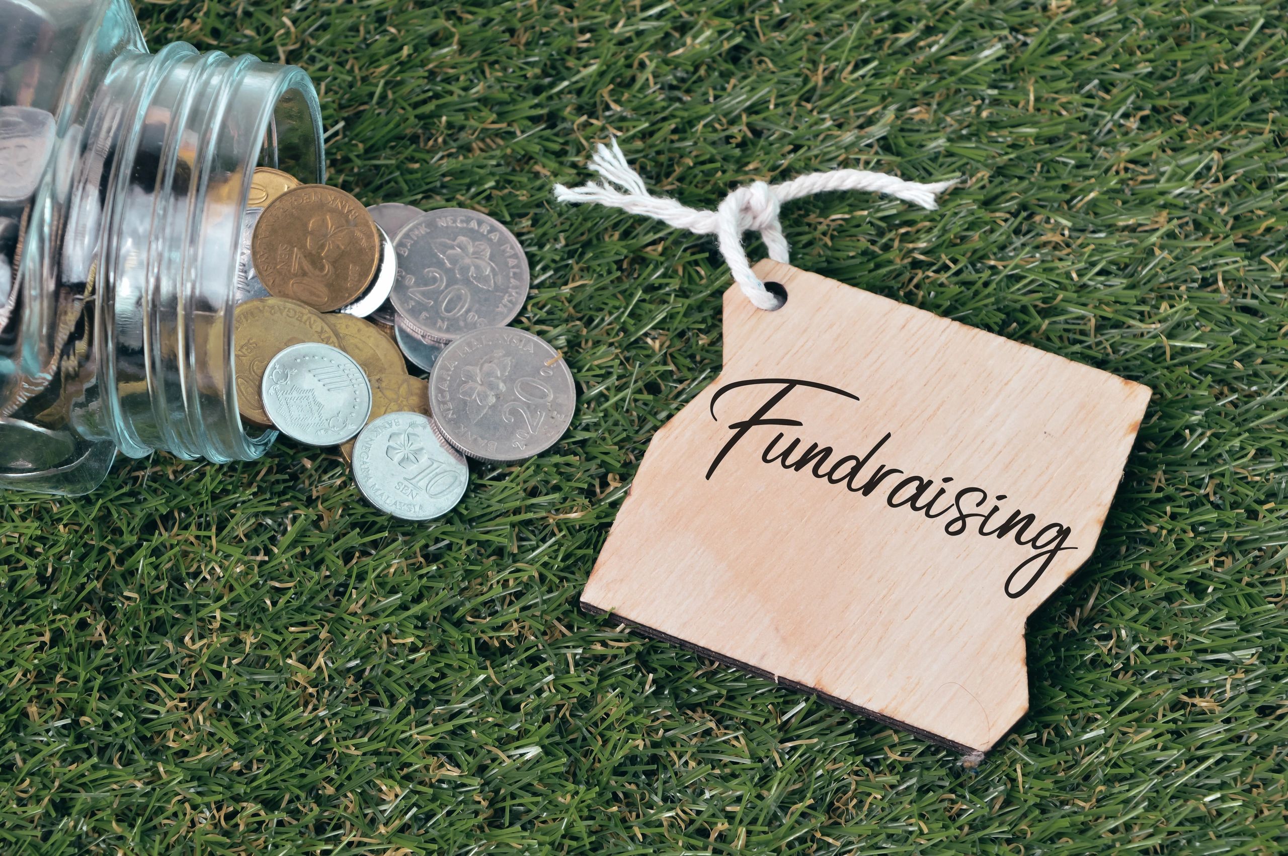 Coins spilling from a jar next to a wooden fundraising tag on grass.