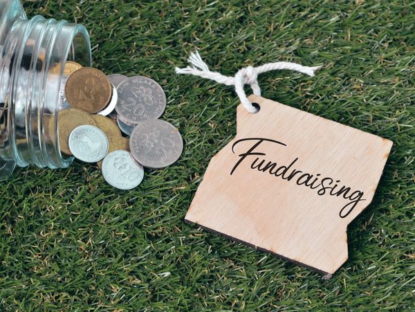 Coins spilled from jar next to wooden tag labeled 'Fundraising' on grass.