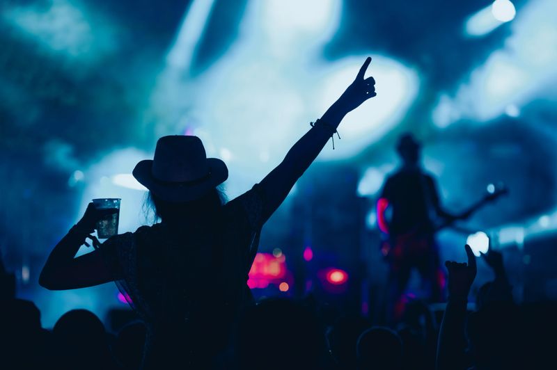 Stage lights against woman with with arms raised while enjoying a concert on a music festival. Crowd with raised hands.
