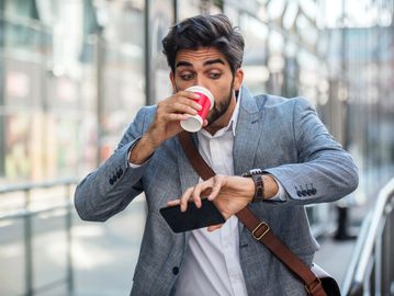 A man in a suit drinking coffee and checking his watch outdoors.