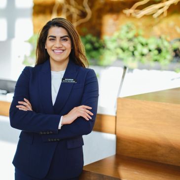 Confident woman in a navy suit smiling with arms crossed in a bright, modern office.