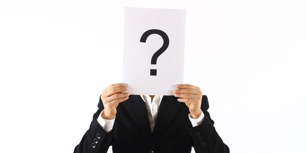 Person in black suit holding paper with question mark covering face.