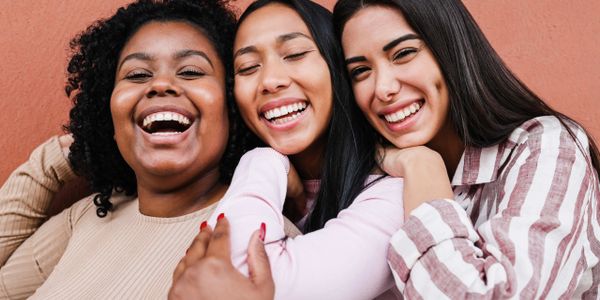Three women laughing and hugging in front of a terracotta wall.