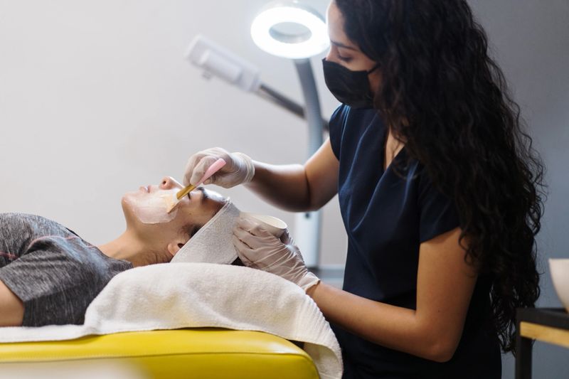 A latin female esthetician applying skin care product to a young woman with a brush.