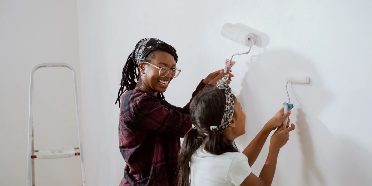 Woman and daughter painting a wall with rollers with paint on their faces and clothes.