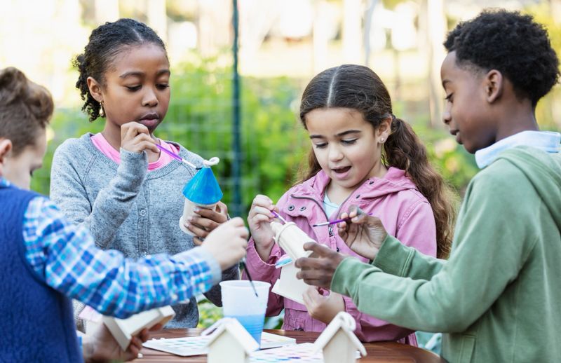 A multi-ethnic group of four children, 7 to 10 years old, doing an art project at a community garden. They are standing outdoors around a table painting little birdhouses or bird feeders. The vegetable garden is out of focus in the background.