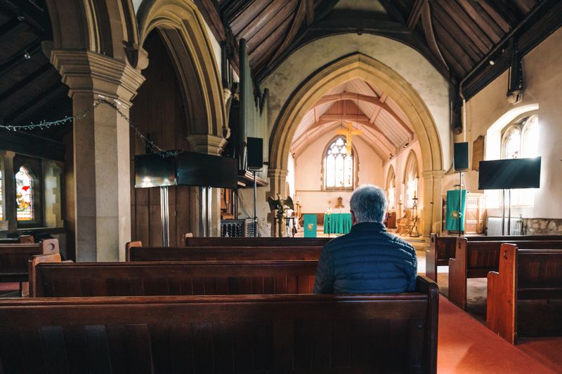 Color image depicting a senior caucasian man in his 60s in quiet prayer inside an old anglican church. The man has his hands clasped together in prayer. The man is alone and has his head bowed in solemn thought. Room for copy space.
