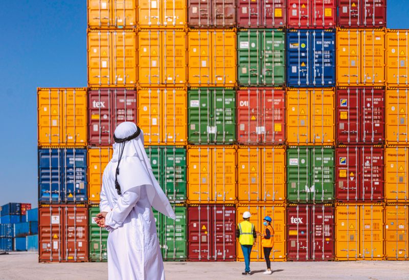 Back side view of young solitary peaceful Emirati Arabian company owner man dressed in traditional clothing kandura, kaffiyeh and agal looking and checking the manufacturer in front of colorful cargo container stacks in shipping port on the large commercial dock. UAE transportation and logistics concept with new normal working lifestyle.