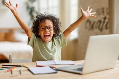 Excited child raising hands at desk with laptop.