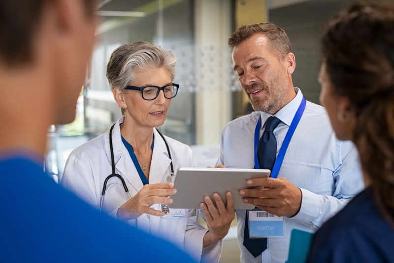 Specialist and woman doctor having a discussion in hospital hallway about new therapy. Senior doctor discussing patient case status with his medical staff after operation. Doctor in conversation showing medical report on digital tablet to pharmaceutical advisor.
