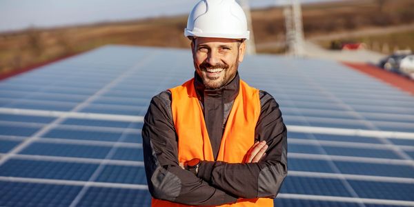 Smiling engineer in safety gear stands confidently on solar panel field.