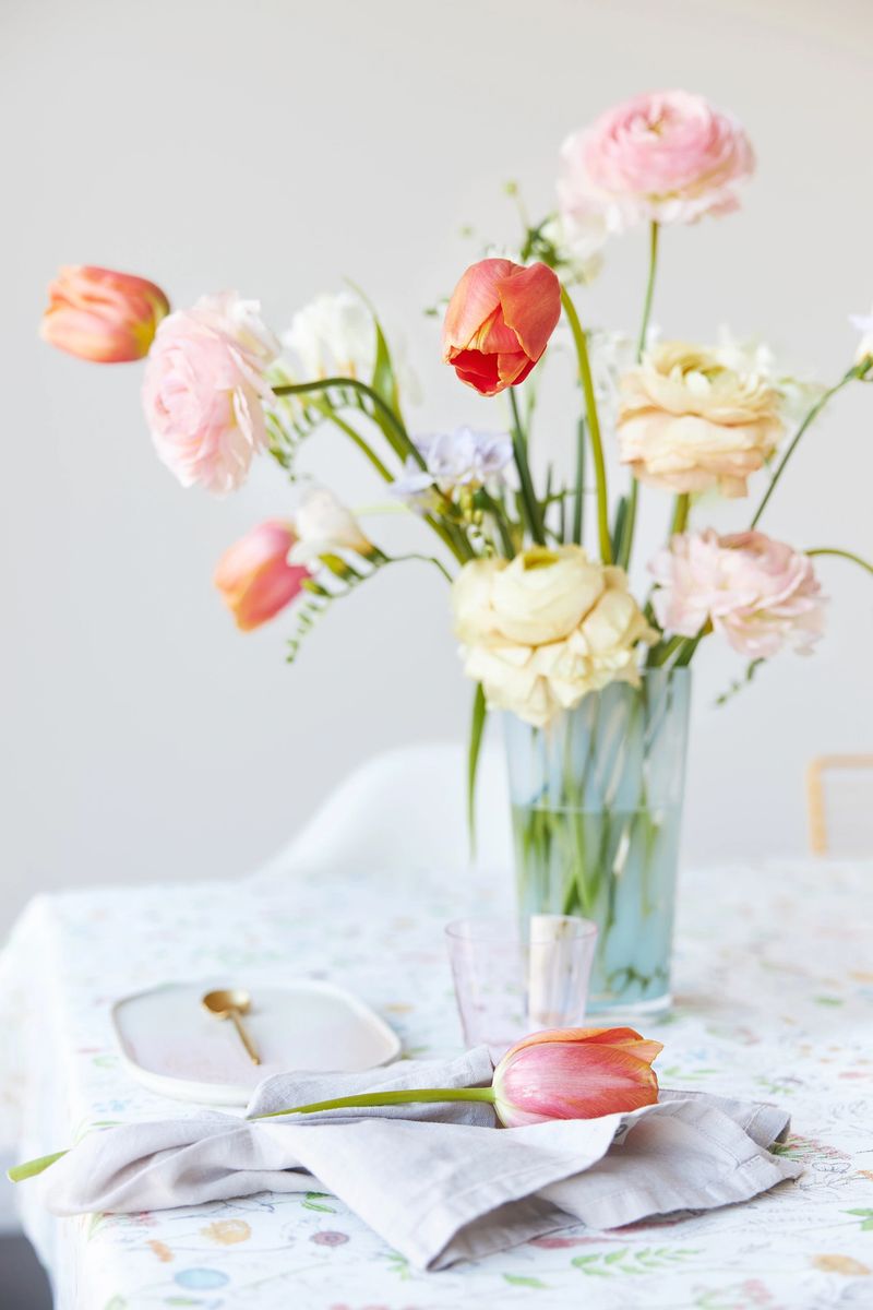 A modern spring bouquet of freesias, ranunculus and tulips in a beautiful vase on a dining table with a floral tablecloth