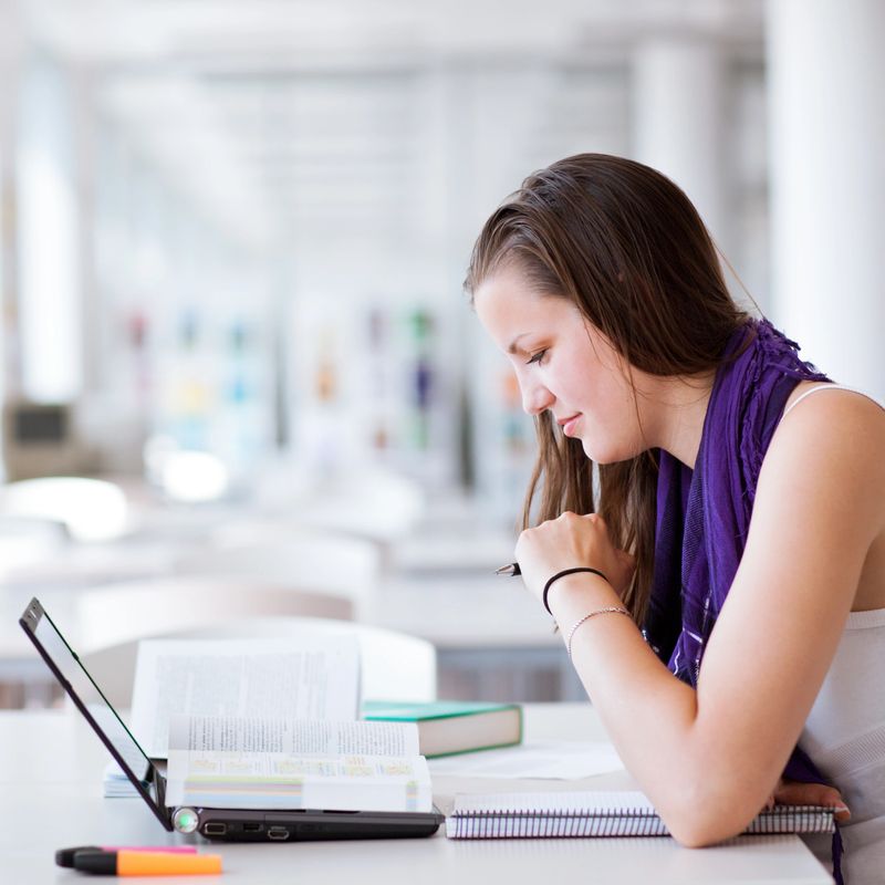 in the library - pretty female student reading and working on her laptop in a college/high school library (color toned image)