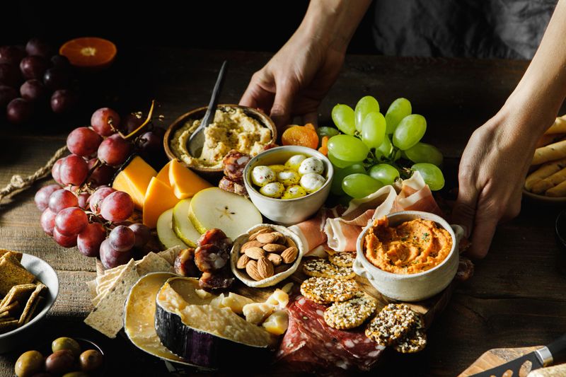 Close-up of a woman serving cheese and meat platter with fruits. Female hands placing mixed fruits with cheese and meat on wooden board.
