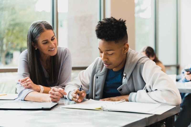 The mid adult female teacher smiles as she helps the teenage boy with his homework.