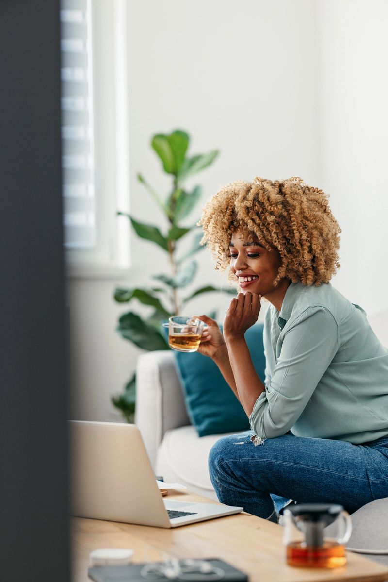 Side view of a smiling Afro-American businesswoman who is sitting on a sofa and holding a glass with a drink while looking at a laptop on the table in front of her. She might be working from home or studying. She is wearing a green shirt and jeans.