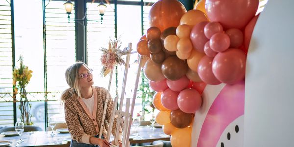 Woman arranging a floral decoration on an easel near colorful balloons indoors.