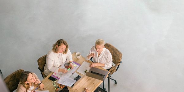 Three women collaborating on a project at a wooden table with laptops and documents.