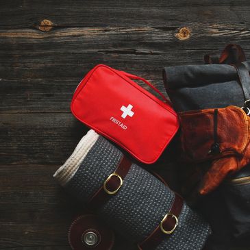 A red first aid kit on a rolled-up blanket and backpack on wooden floor.