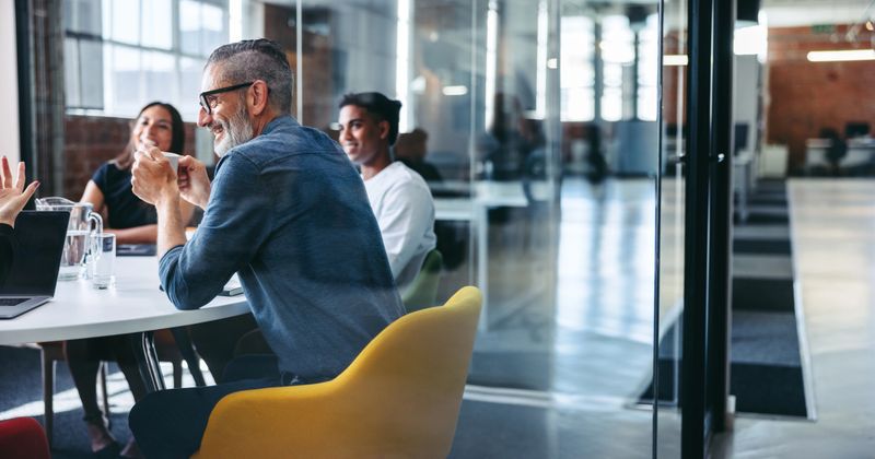 Cheerful mature businessman attending a meeting with his colleagues in an office. Experienced businessman smiling while sitting with his team in a meeting room. Creative businesspeople working together.