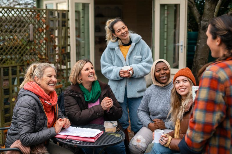 Volunteers sitting outdoors wearing warm casual clothing on a sunny cold winters day. They are resting and having a tea break from working on a community farm, looking after crops and performing other sustainable and environmentally friendly tasks. They are laughing and talking together, drinking hot drinks.