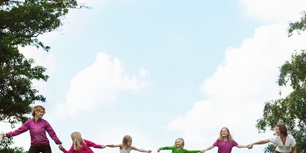Two adults and four children hold hands, playing outdoors on a grassy hill under a blue sky.