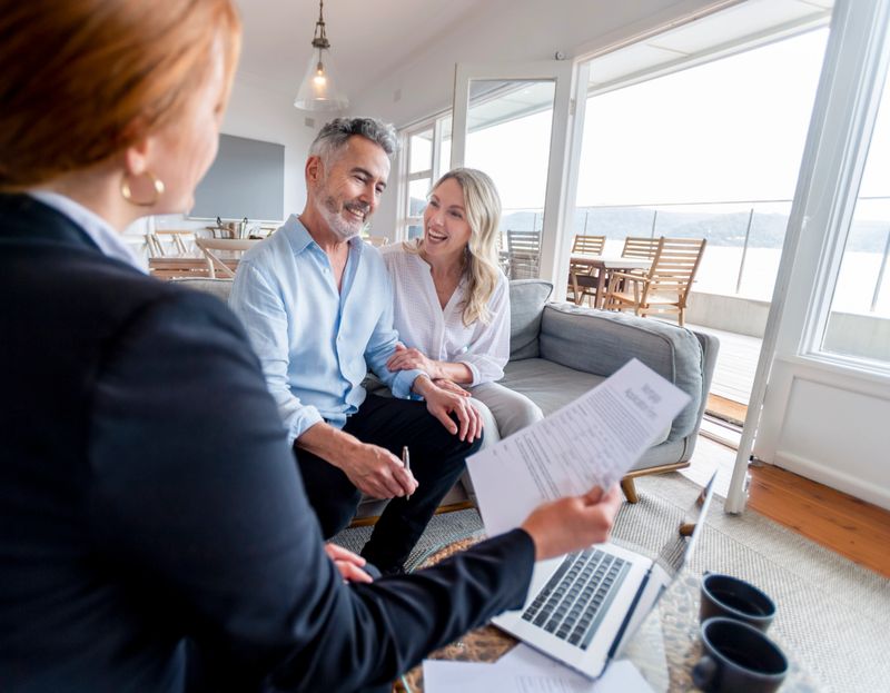 Happy mature couple meeting investments and financial advisor at home. They are happy and smiling sitting in the living room. The advisor is holding a document. There is a laptop on the table