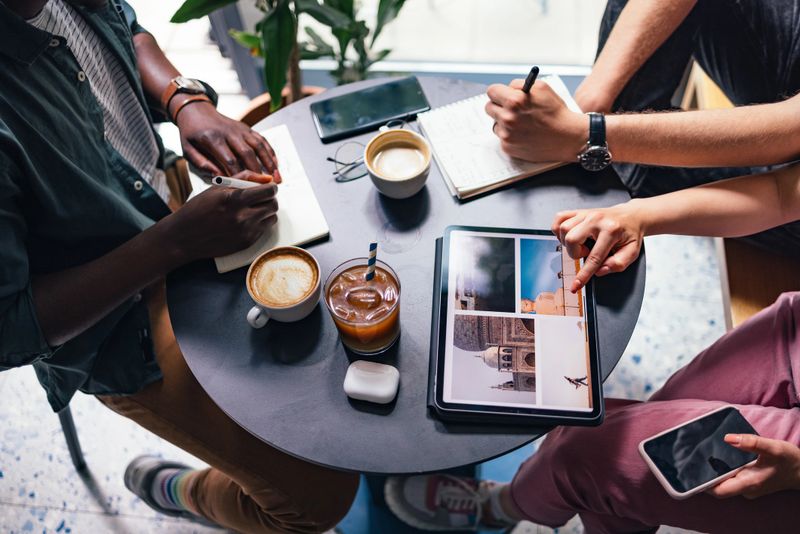High angle view of unrecognizable multi ethnic group of friends sitting in cafe and looking at travel photos on a digital tablet while drinking coffee and writing notes in notebook.