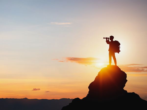 Silhouette of a hiker with a backpack using binoculars on a mountain at sunset.