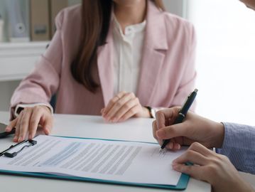 A person signing a document with a woman in a pink blazer.