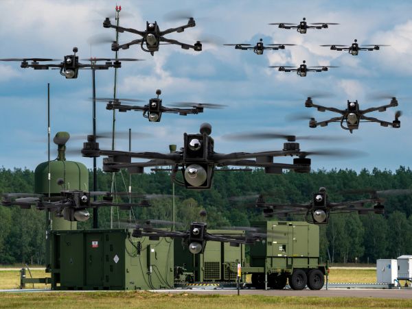 Multiple drones flying above military equipment in an open field.
