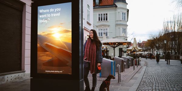 A woman with a dog stands near a travel advertisement in a city street.