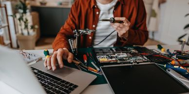 Person repairing electronics with tools while referencing a laptop on a workbench.