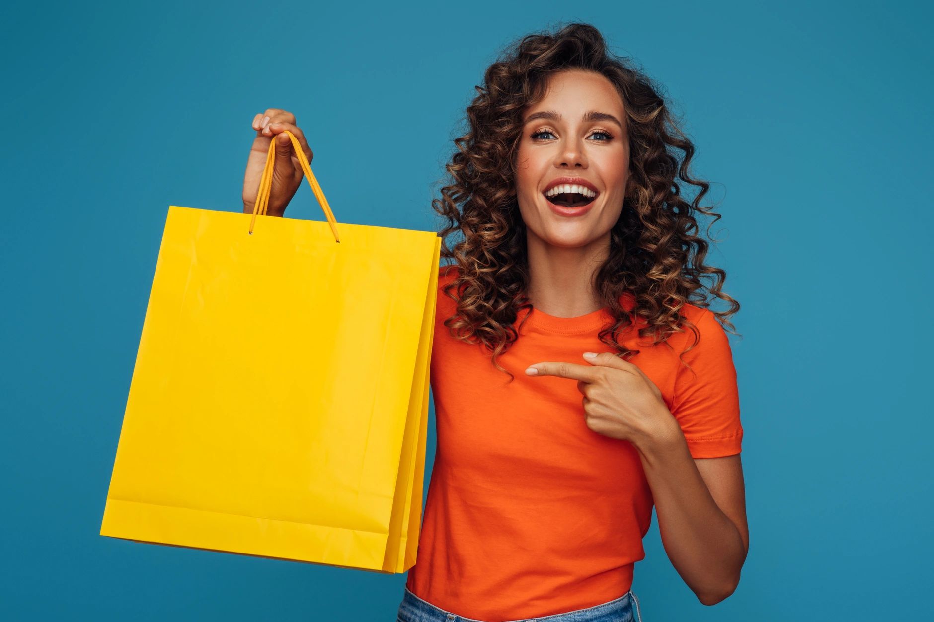 A smiling woman wearing a orange t-shirt with yellow shopping bags and happiness-wellbeing product.