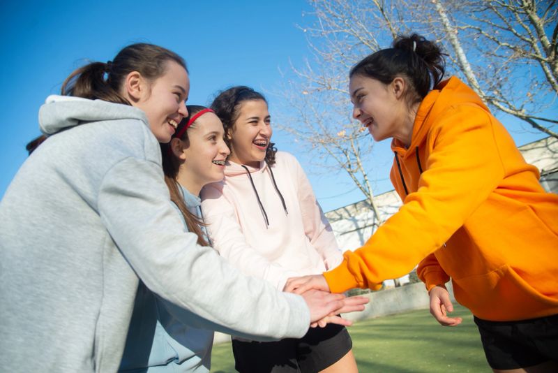 Happy girls of football team enjoying success.Four smiling girls standing on green field after training holding hands, talking of achievements and progress in game.Team sport, active lifestyle concept