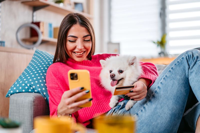 Beautiful young woman sitting on the sofa in the living room with her dog and shopping online using her phone.