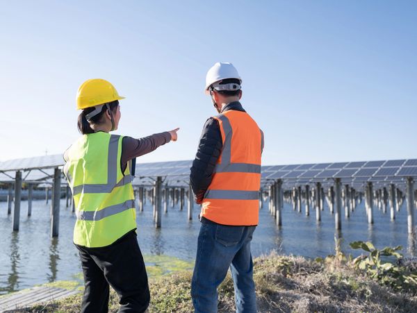 Two engineers in safety gear inspecting solar panels over water.