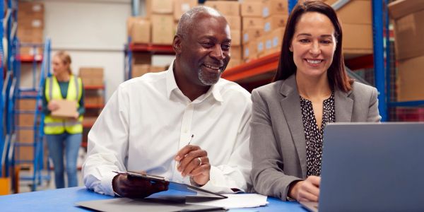 Two colleagues smiling while working on a laptop in a warehouse.
