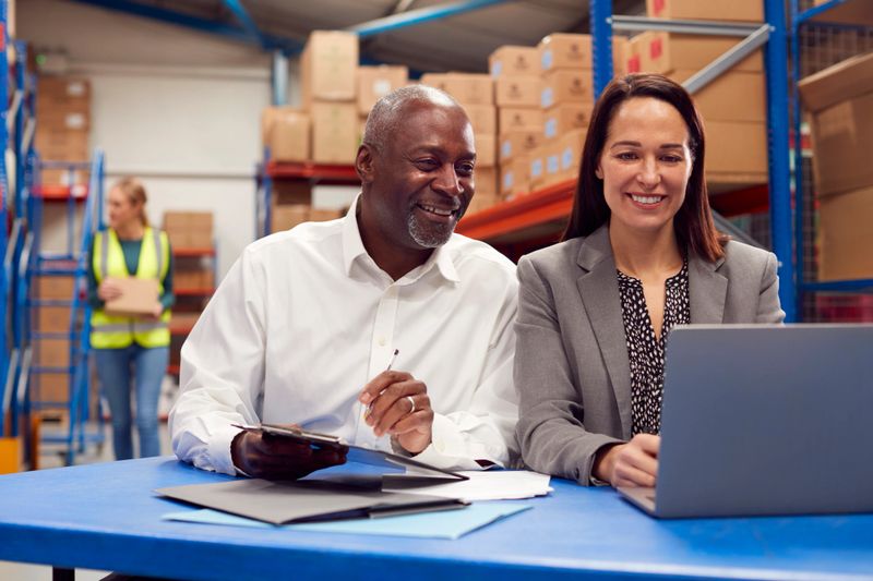 Male And Female Team Leaders Working On Laptop In Warehouse With Workers In Background