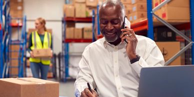 Man in warehouse office on phone, signing documents with a laptop nearby.