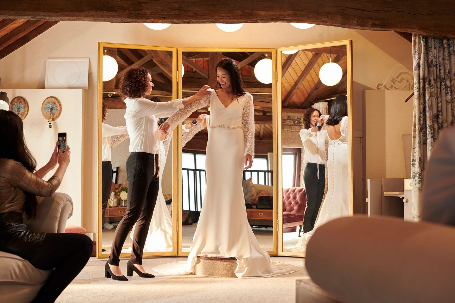 Bride trying on a wedding dress with a tailor's assistance and a friend taking a photo.