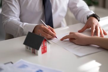 A person signing a document with a small house model nearby.
