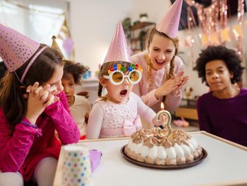 Children wearing party hats celebrate a girl's 3rd birthday with a cake and candles.