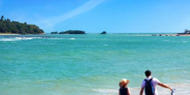 A couple walking barefoot along a sunny beach with turquoise water.