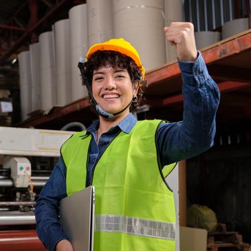 Smiling female worker in safety gear celebrating success in an industrial setting.