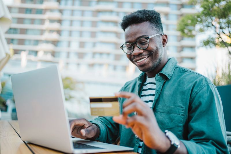 Shot of young man doing shopping online with laptop and payment by credit card. Cheerful man using laptop computer and credit card in a sidewalk cafe.