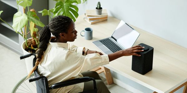 Woman in wheelchair adjusts speaker on desk with laptop and plants.